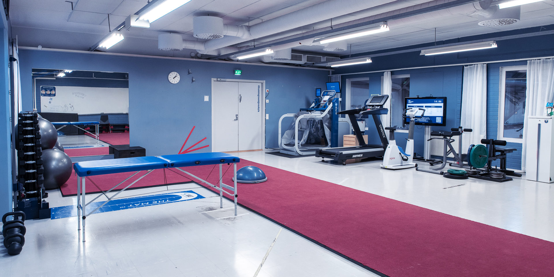 Kuva tyhjästä Training Roomista, jossa näkyy punainen pitkä jumppamatto, hierontapöytä, jumppapalloja, juoksumatto ja muita harjoituslaitteita.