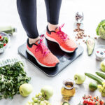 Urheilijan naisen jalat näkyvissä lenkkikengät jalassa seisoen vaa'alla. Ympärillä maassa terveellisiä ruokia, hedelmiä ja vihanneksia.
